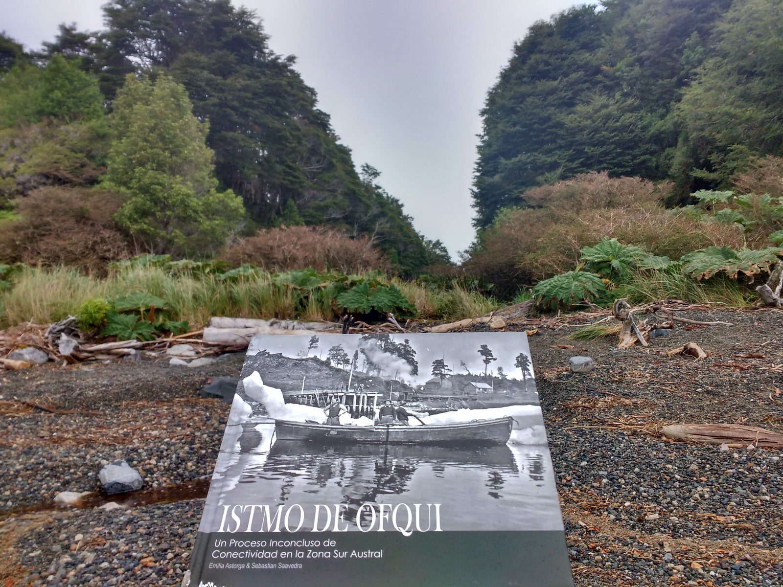 Portada del libro «Istmo de Ofqui» sobre una playa de grava, rodeada de bosque